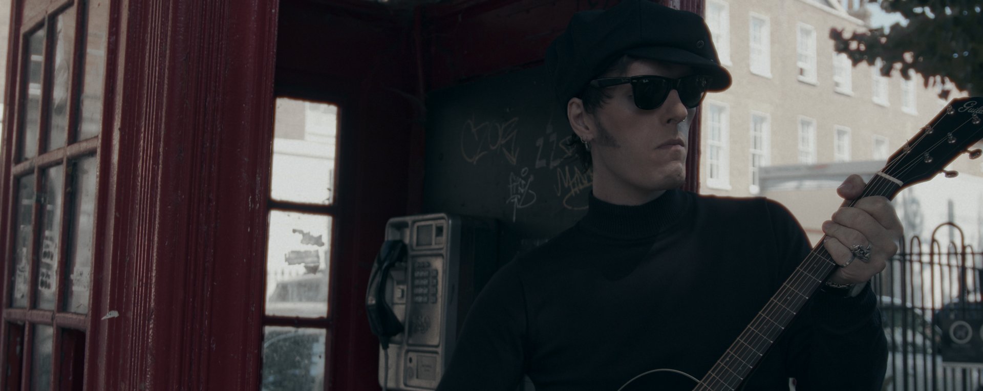 With an acoustic guitar beside a red phone box on a London street.