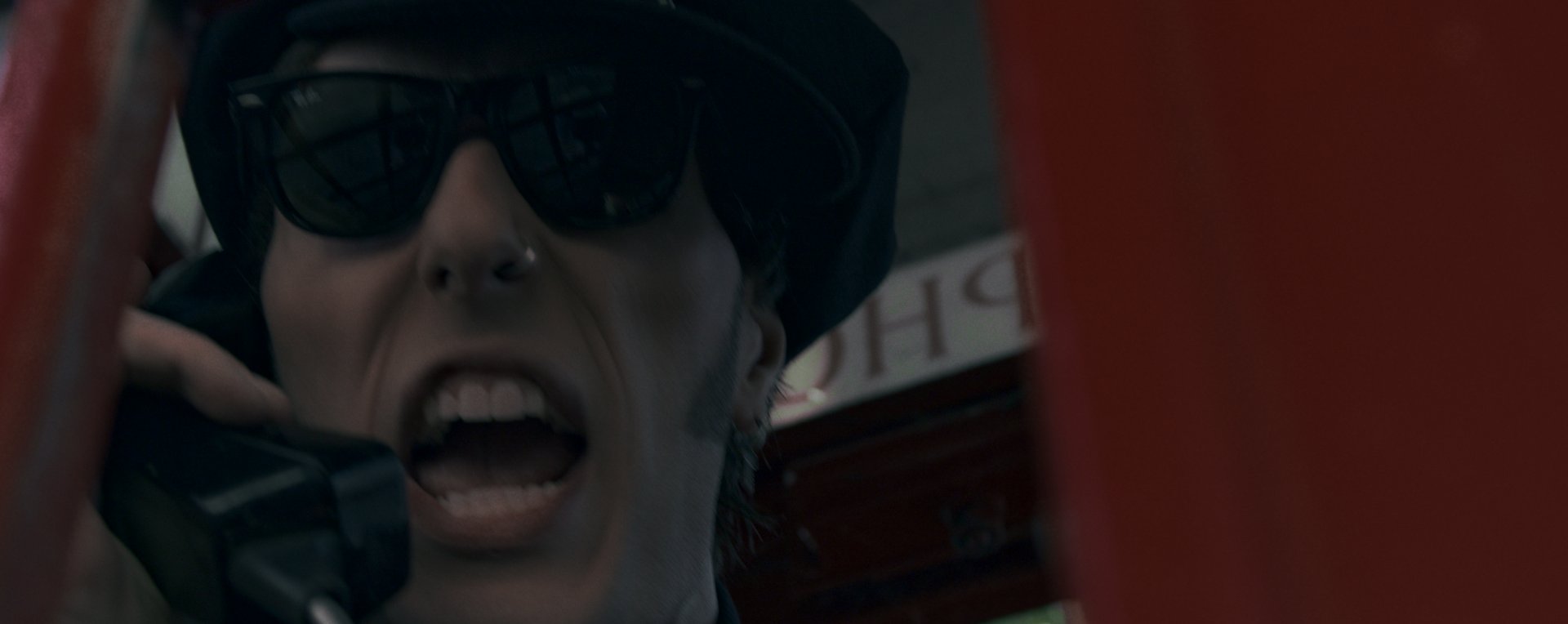 Close on his face mid-shout against the red of the phone box.