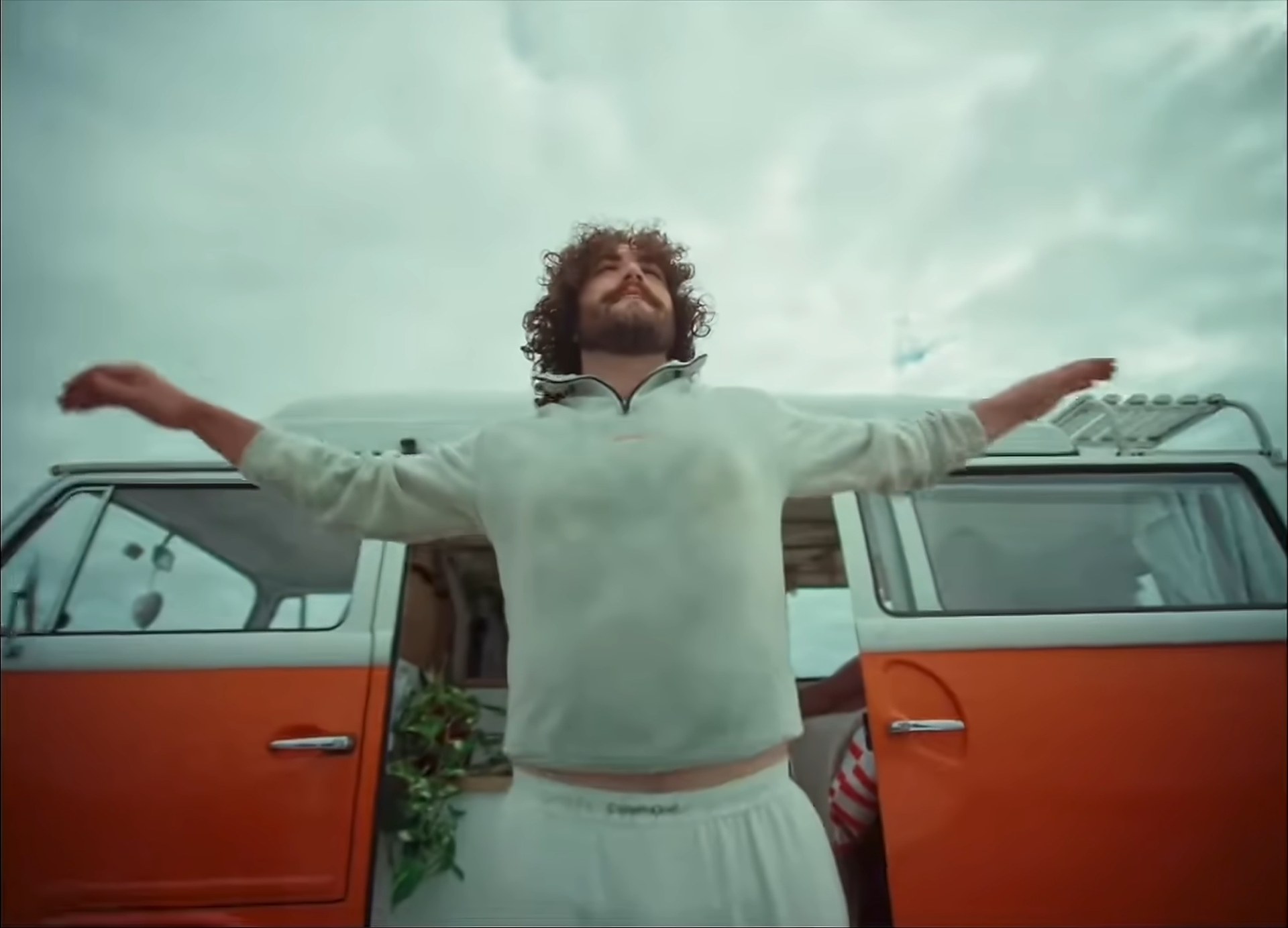 A man with his arms spread wide against the open doors of an orange camper van, under a big sky.