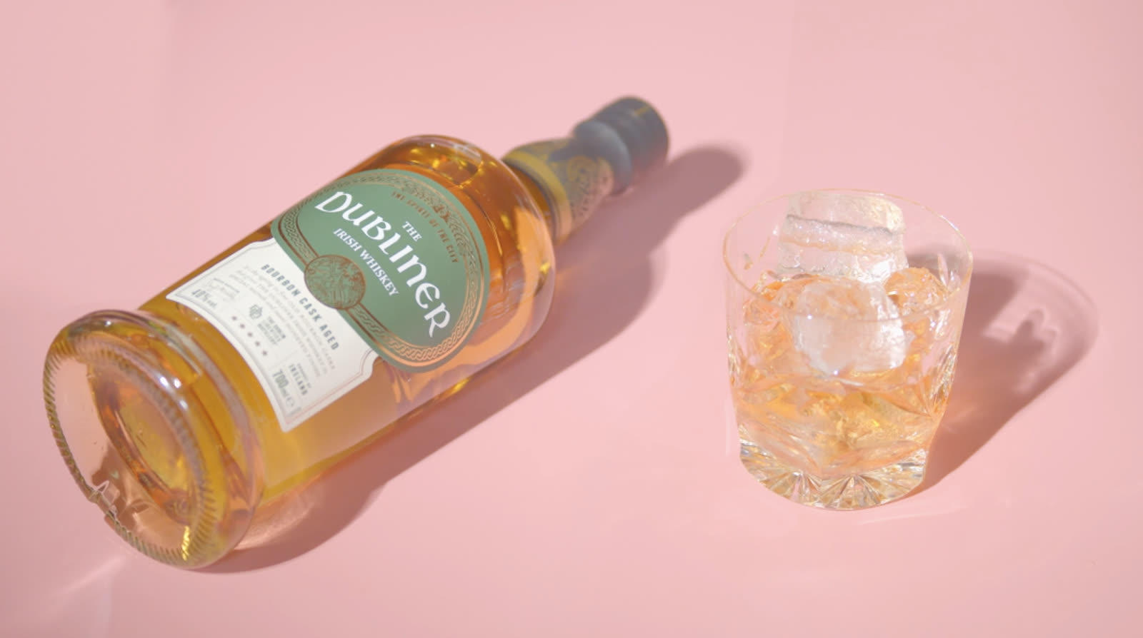 A bottle of The Dubliner Irish whiskey lying beside a cut-glass tumbler on a pink ground.