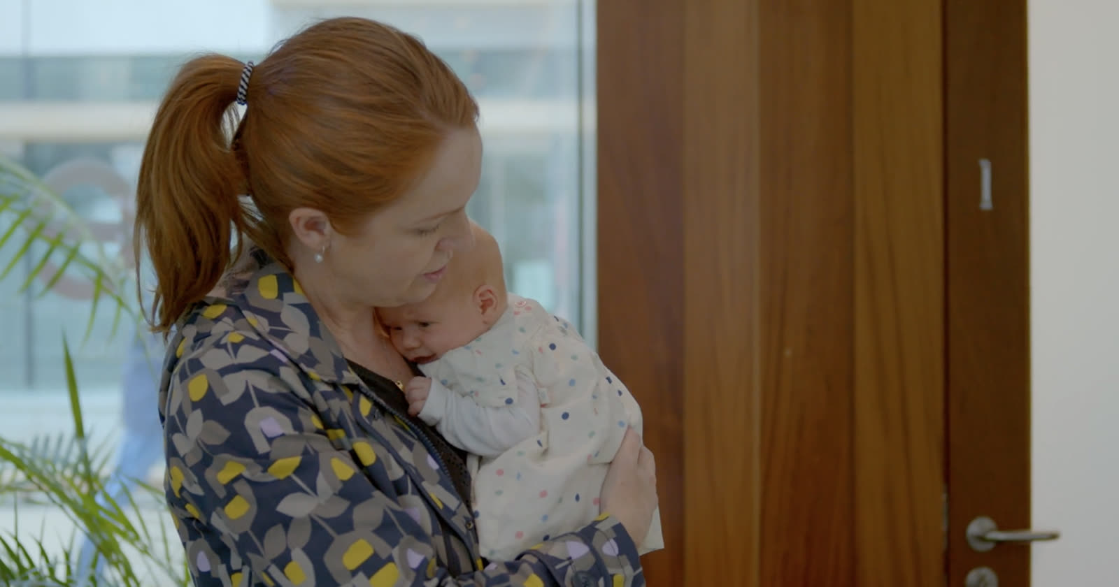 A mother holding a newborn close, standing by a bright window.