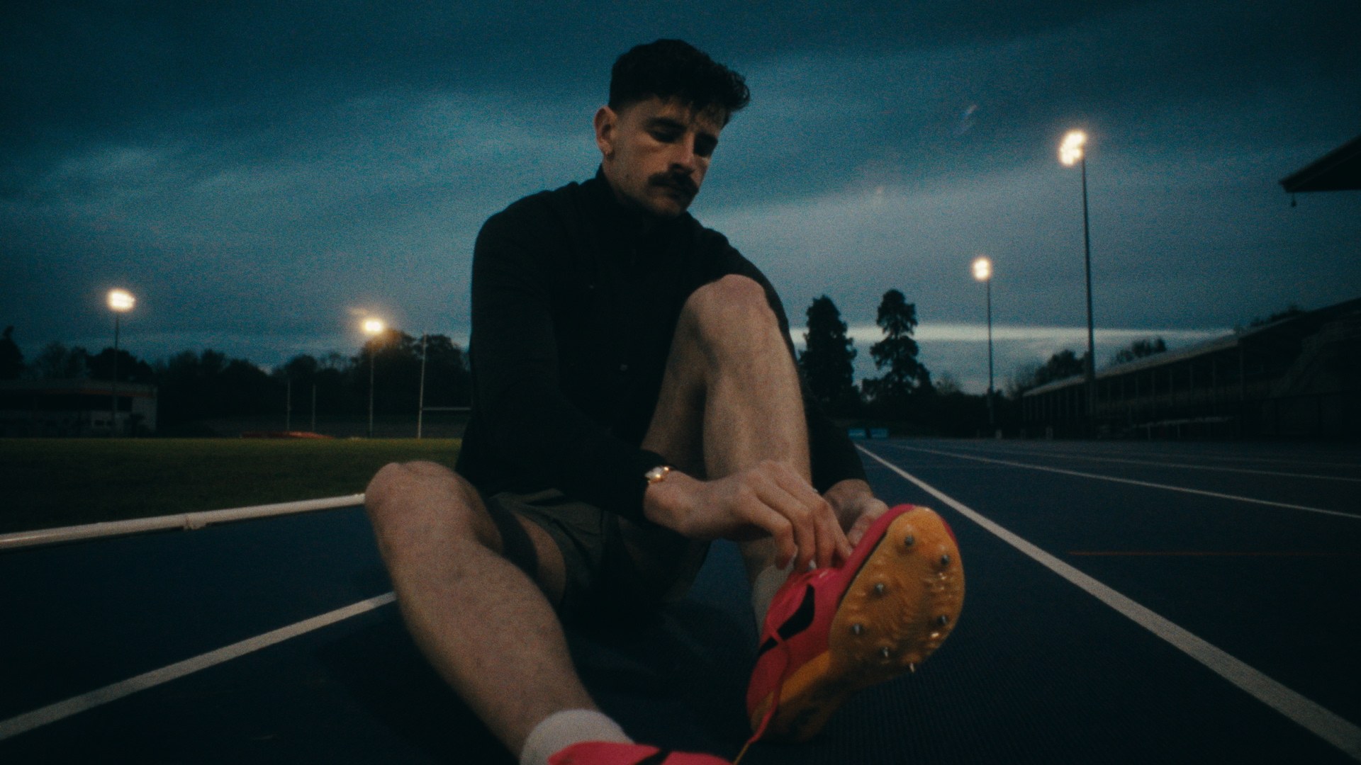 A runner sitting on the track at dusk, floodlights behind him.