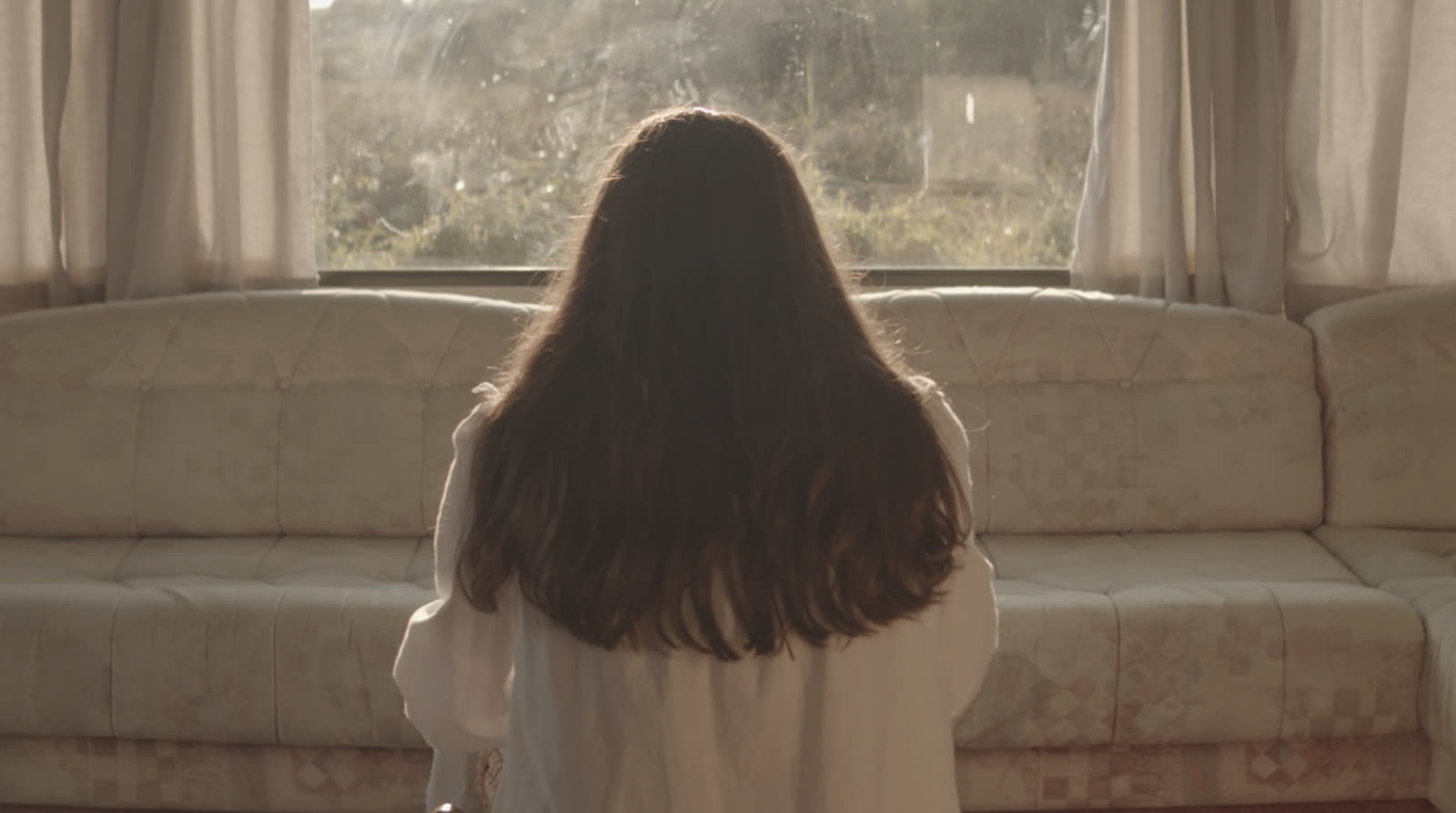 A girl seen from behind, facing a bright caravan window in soft morning light.