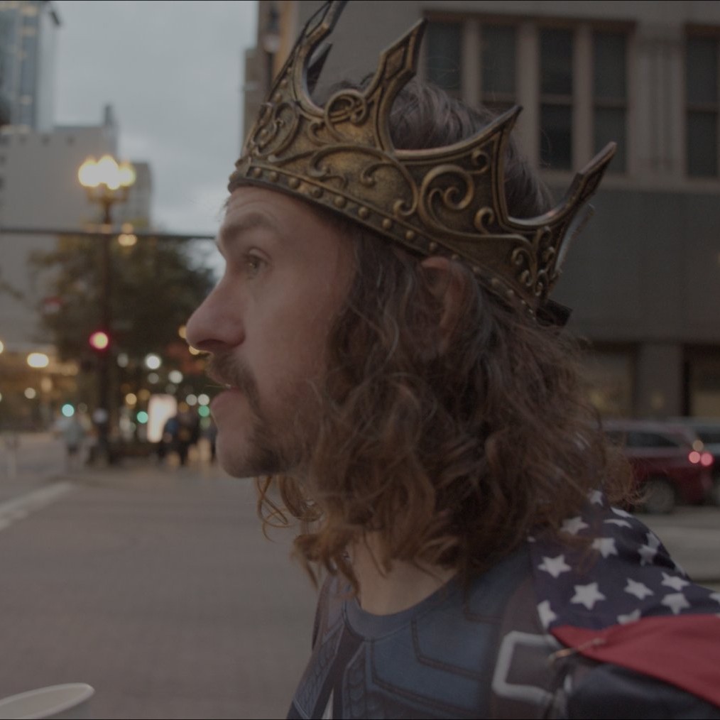 In a crown and a flag cape, in profile on an empty Chicago street.