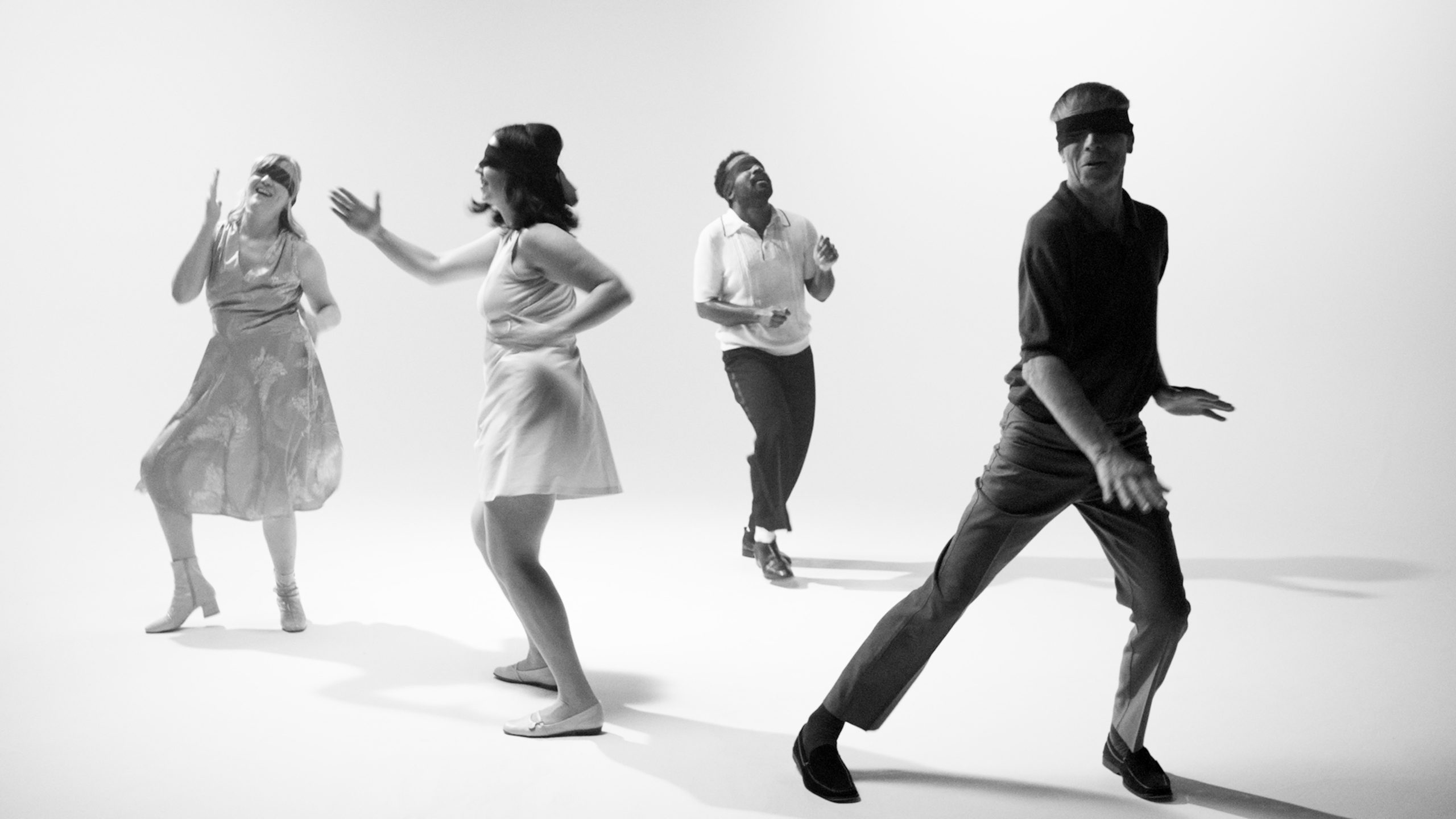 Four blindfolded dancers doing the twist in a bright white void, caught mid-step.