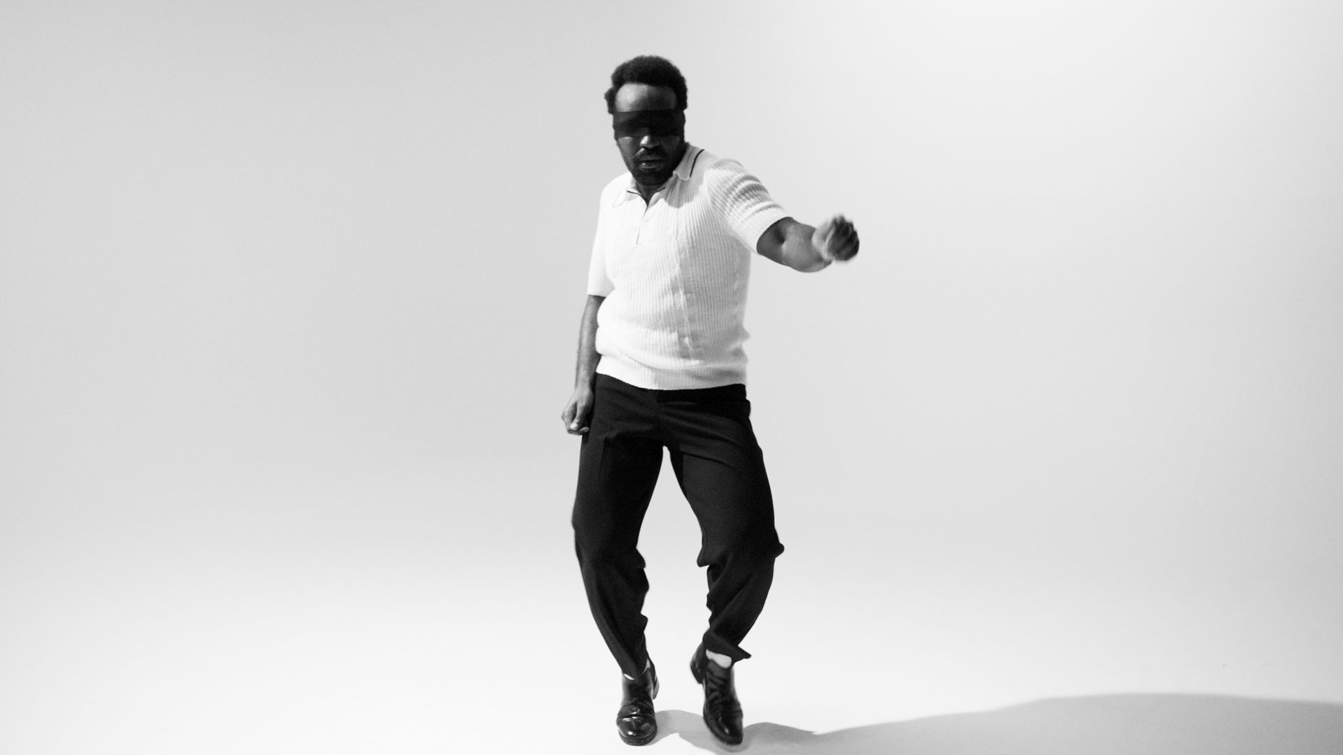 A blindfolded dancer alone in the white, arm extended, both feet off the floor.