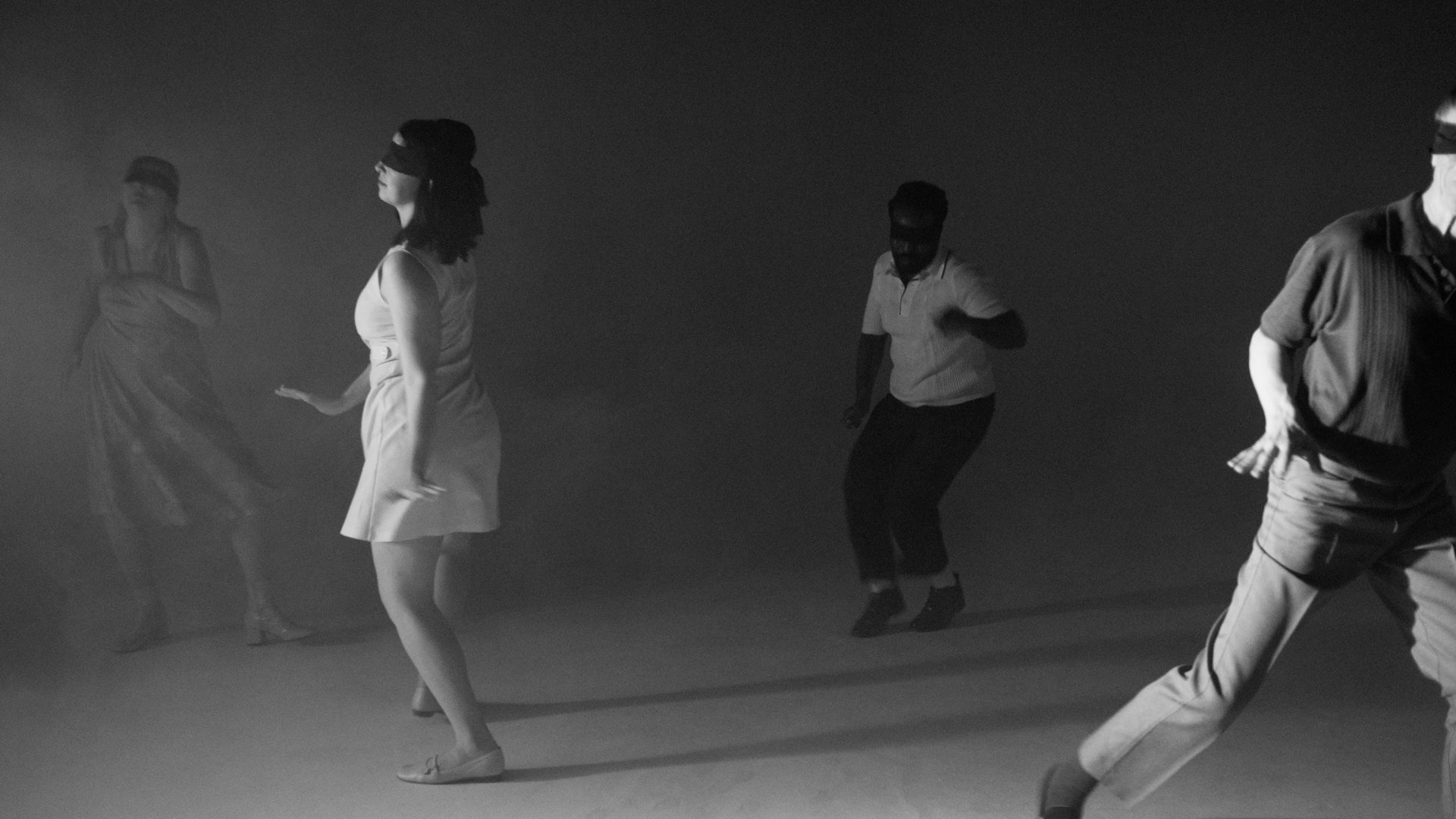 The same four dancers, now in smoke and low light, the white void gone dark.