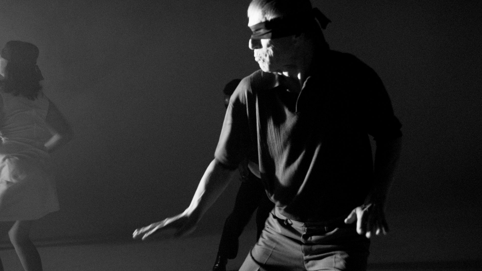 A blindfolded dancer close up in the dark, lit hard from the side, hands low.